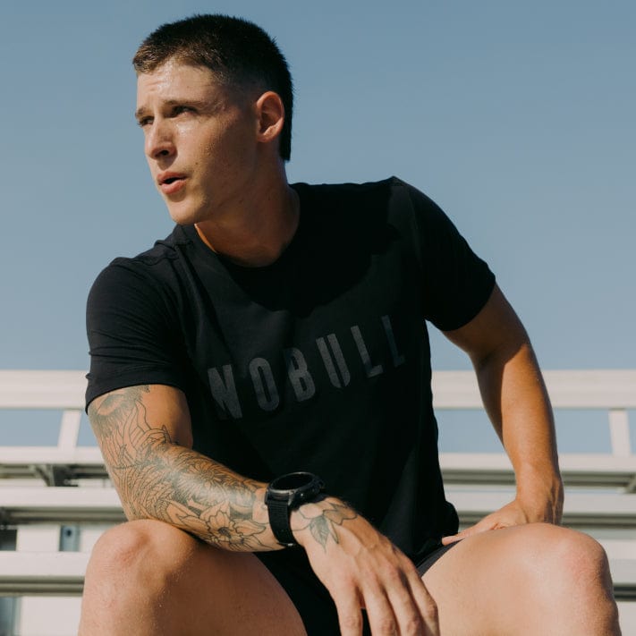 Man wearing a black t-shirt with 'NOBULL' logo, sitting outdoors against a clear sky.